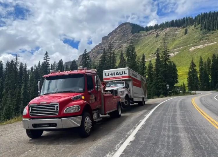 A red tow truck is towing a u-haul trailer down a mountain road.