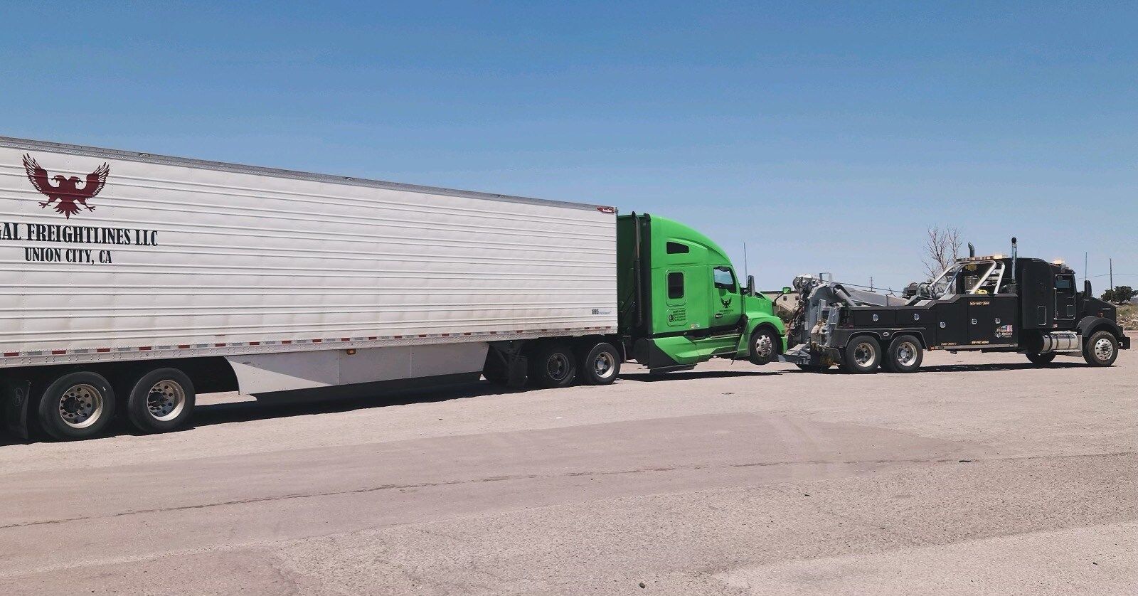 A green semi truck is being towed by a black tow truck.