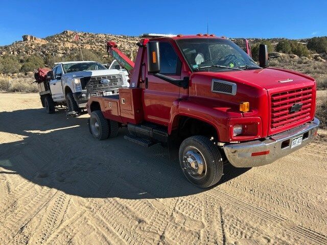 A red tow truck is parked next to a white truck on a dirt road.