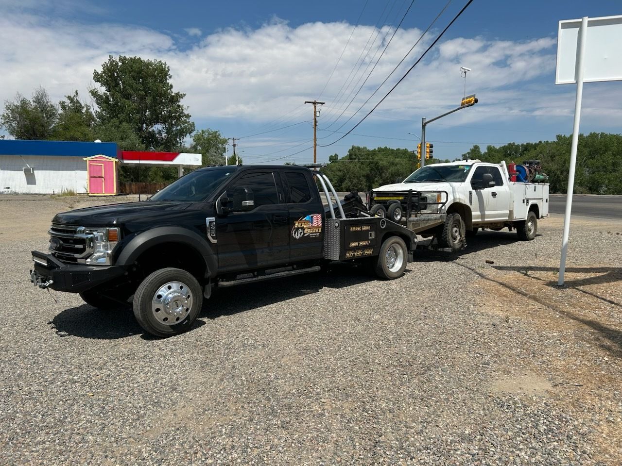 A black truck is towing a white truck in a gravel lot.