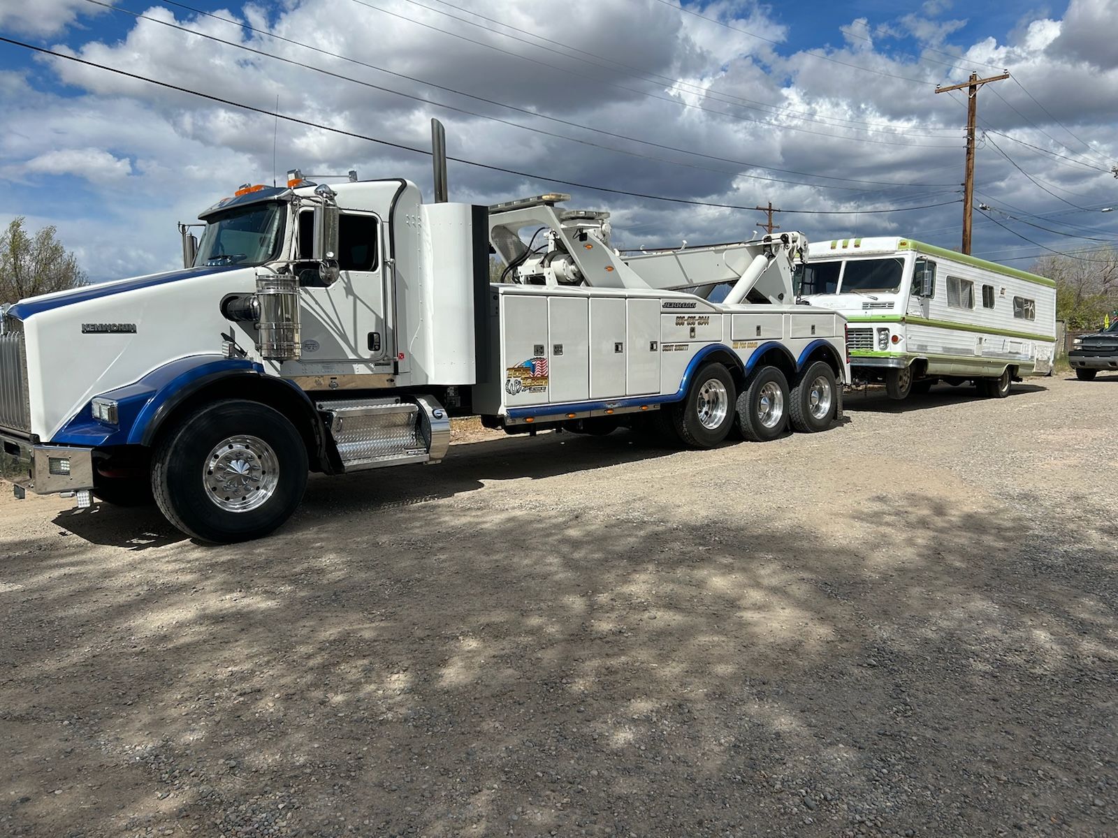 A white tow truck is towing a trailer in a parking lot.