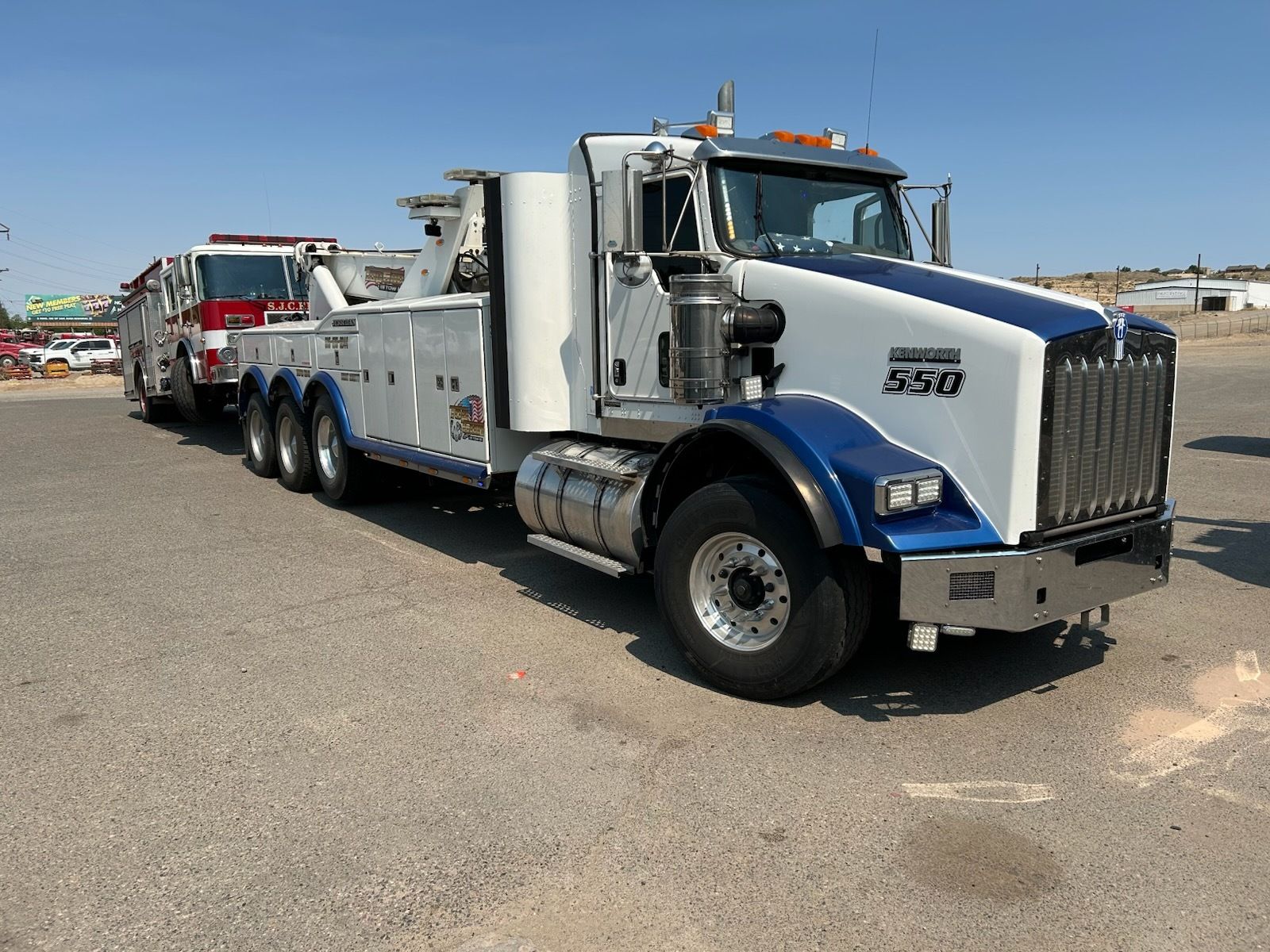 A white and blue tow truck is parked in a parking lot next to a fire truck.