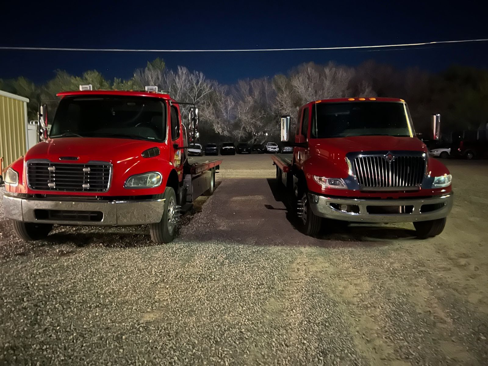 Two red tow trucks are parked next to each other in a gravel lot at night.