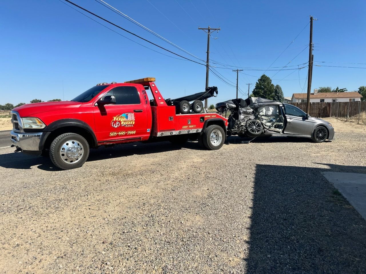 Red tow truck towing a severely damaged silver car on a gravel road under a clear blue sky.