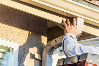 Person installing a gutter. Beige siding, gray gutter, outdoors.