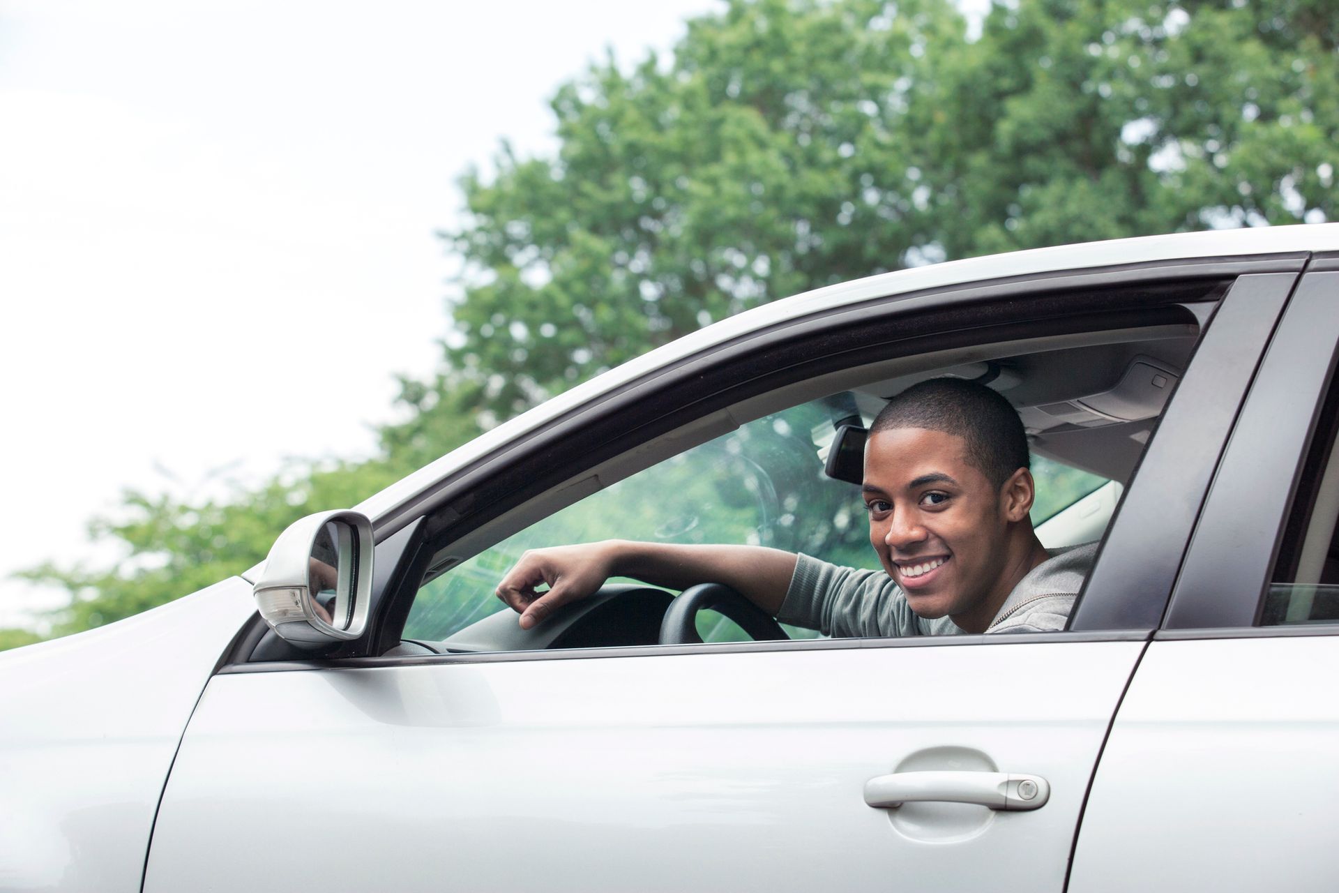 Teenager drives a silver car, with a blurry tree and white sky in the background.