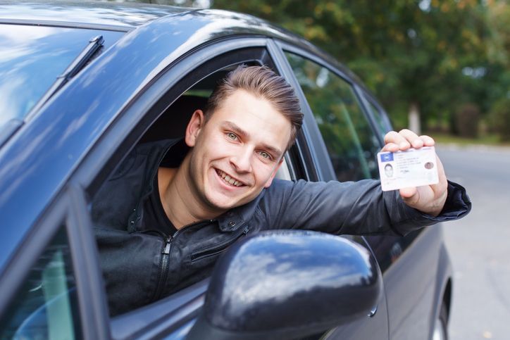 Portrait of a young man leaning out from the driver’s seat window as he shows his driving license.>