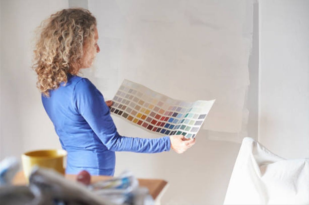 A person holds a colour swatch while choosing paint for a partially painted interior wall.