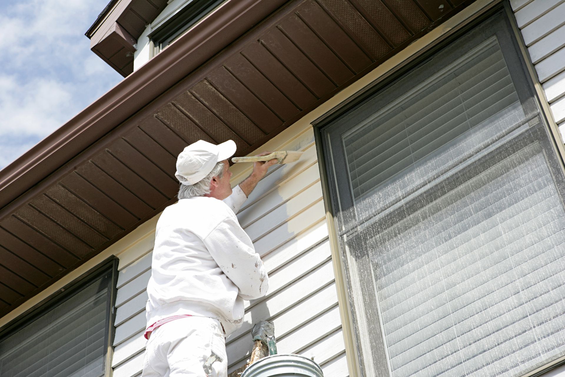 Residential painter in white uniform paints the exterior of a house with white paint.