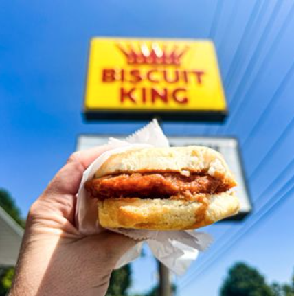 Hand holding a Biscuit King biscuit sandwich under a yellow sign against a blue sky.