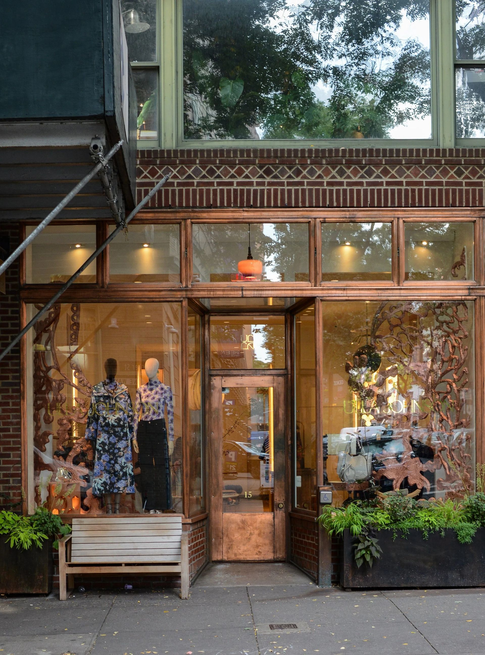 Storefront with copper-colored trim and glass windows; plants and bench in front, mannequins inside.