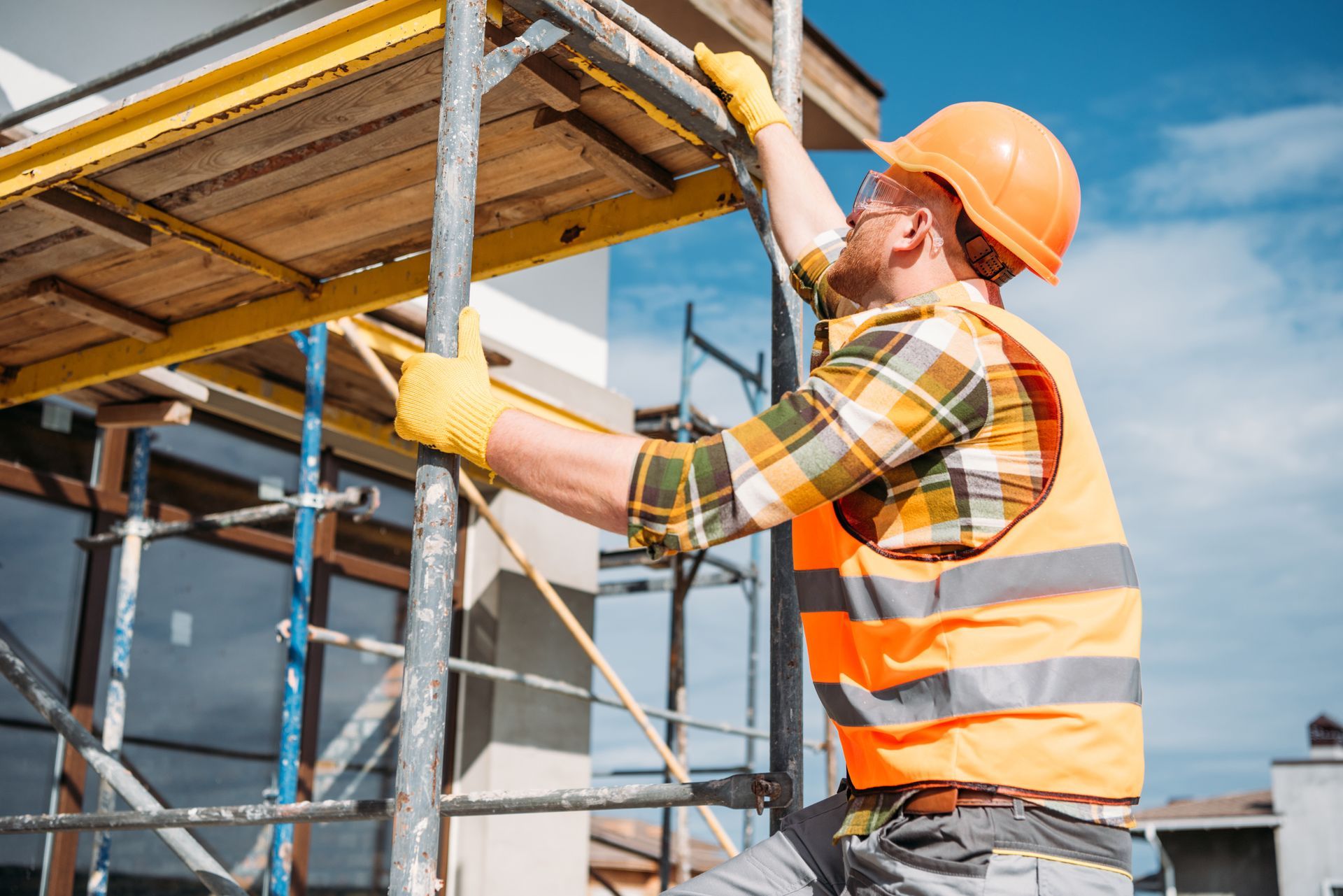 A builder, wearing an orange helmet, is climbing on scaffolding at a construction site.