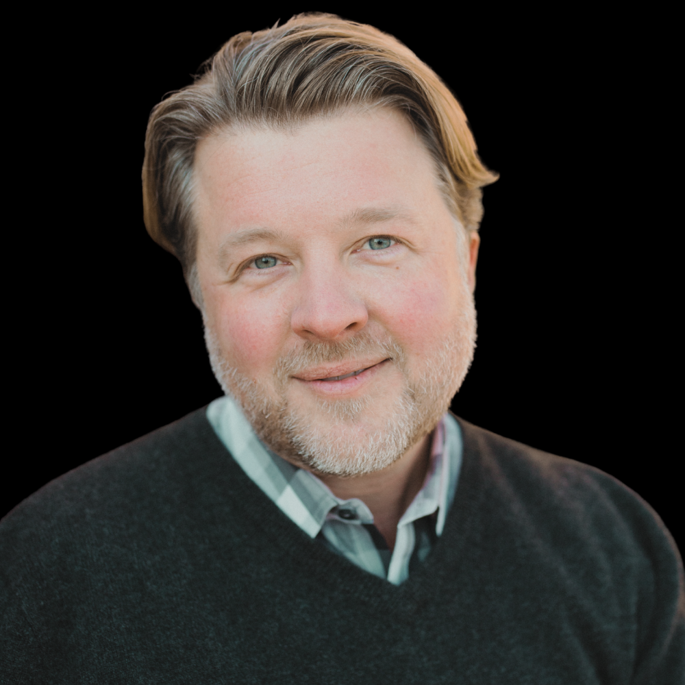 White man with light brown hair and a grey sweater smiling against a metal staircase