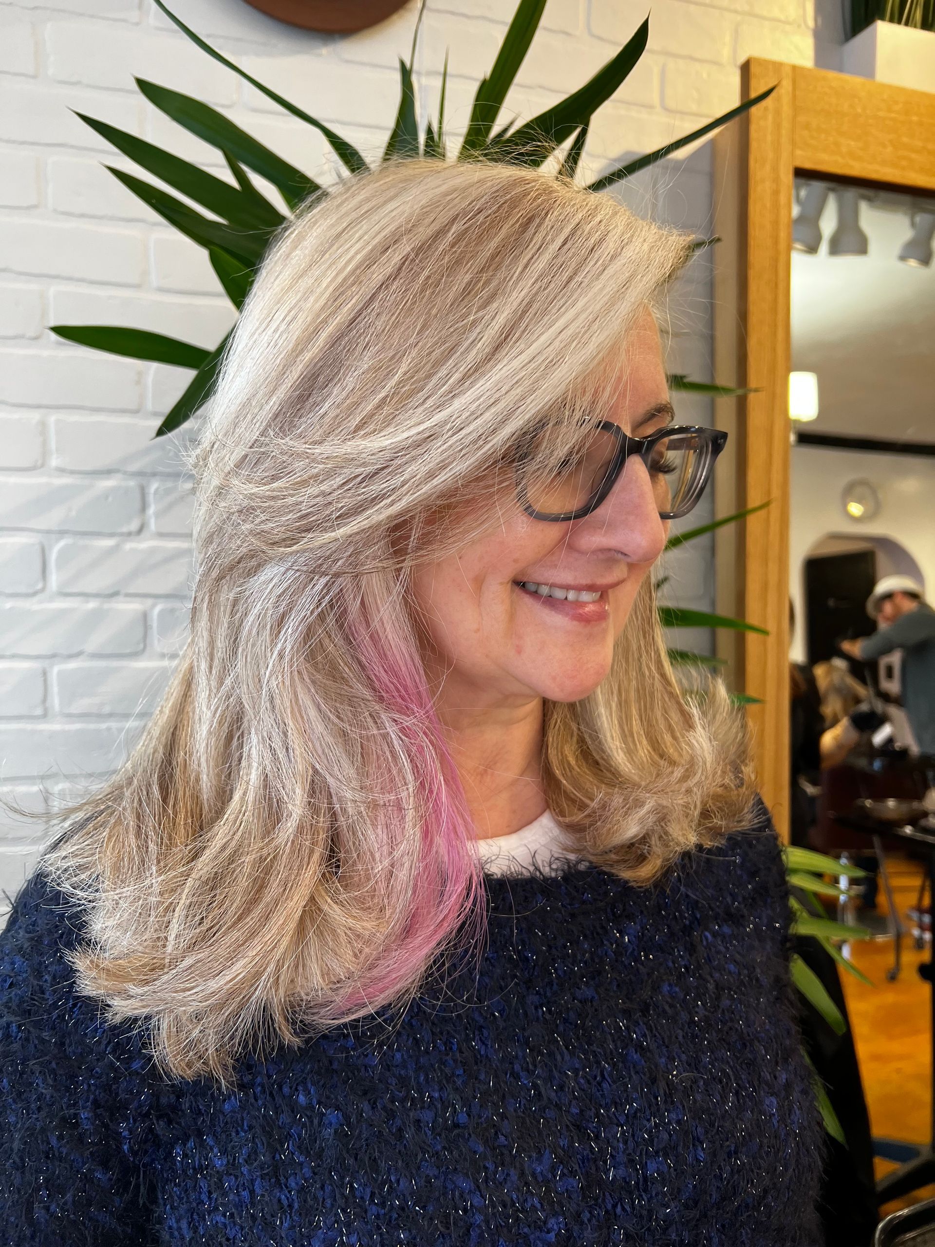A woman with blonde hair and pink streaks is wearing glasses and a blue sweater