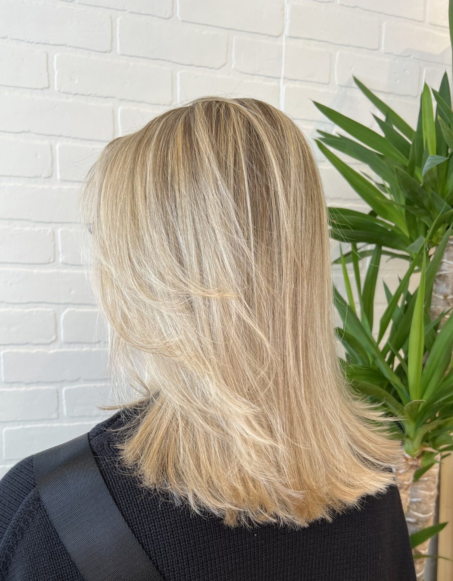 A woman with blonde hair is wearing a black sweater and standing next to a plant.