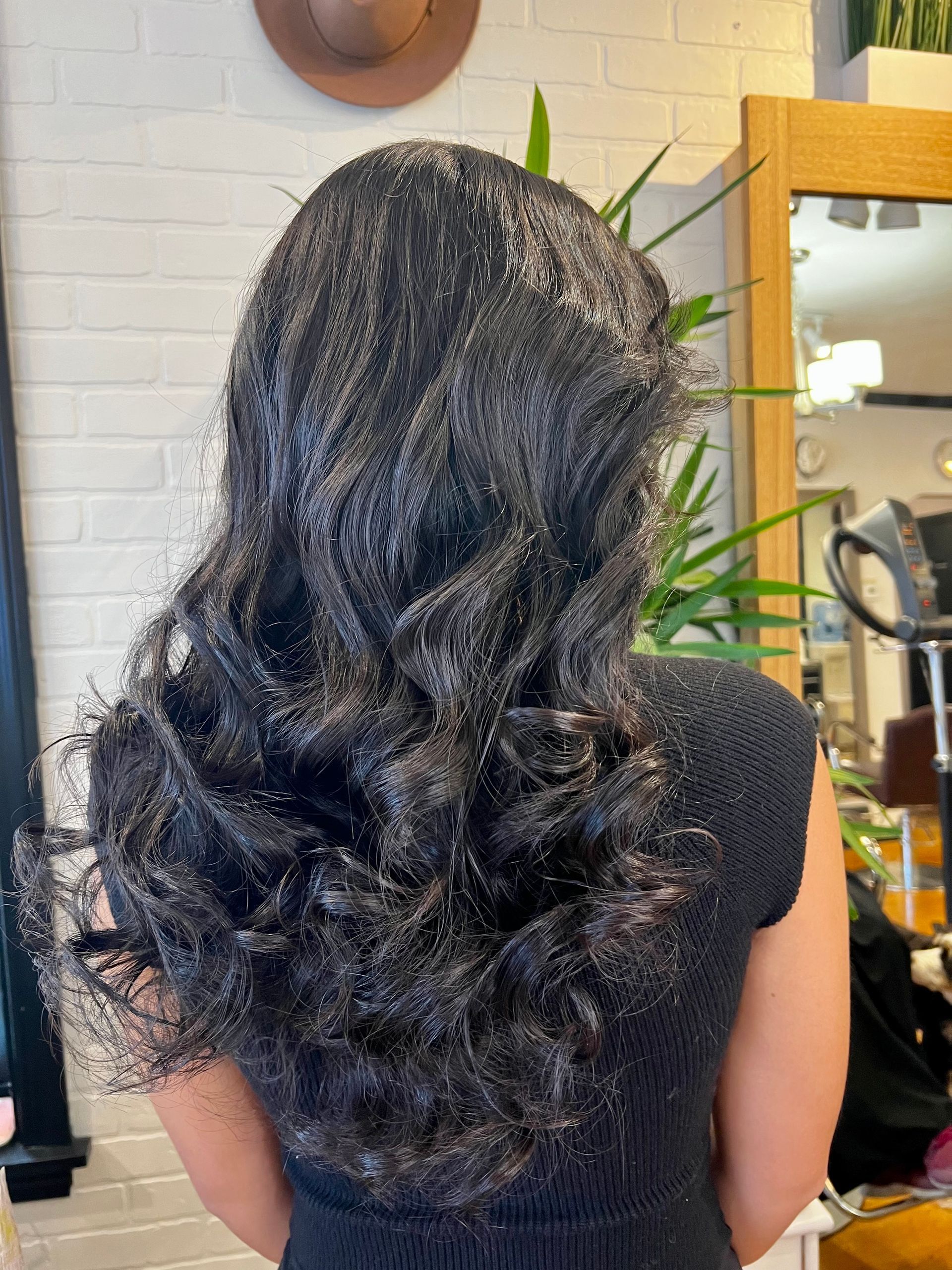 A woman with long black hair is standing in front of a mirror in a salon.