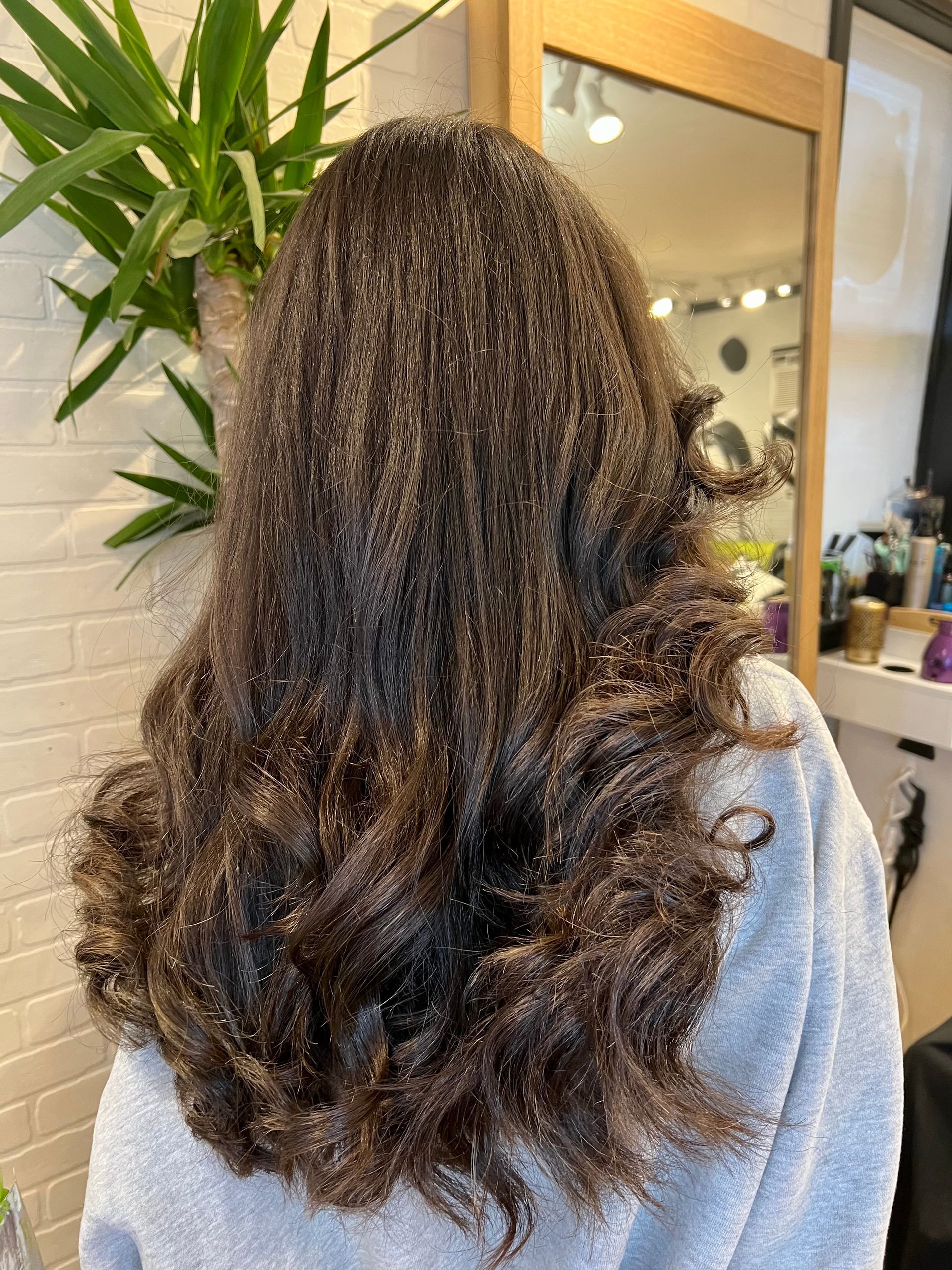 A woman with long curly hair is standing in front of a mirror in a salon.