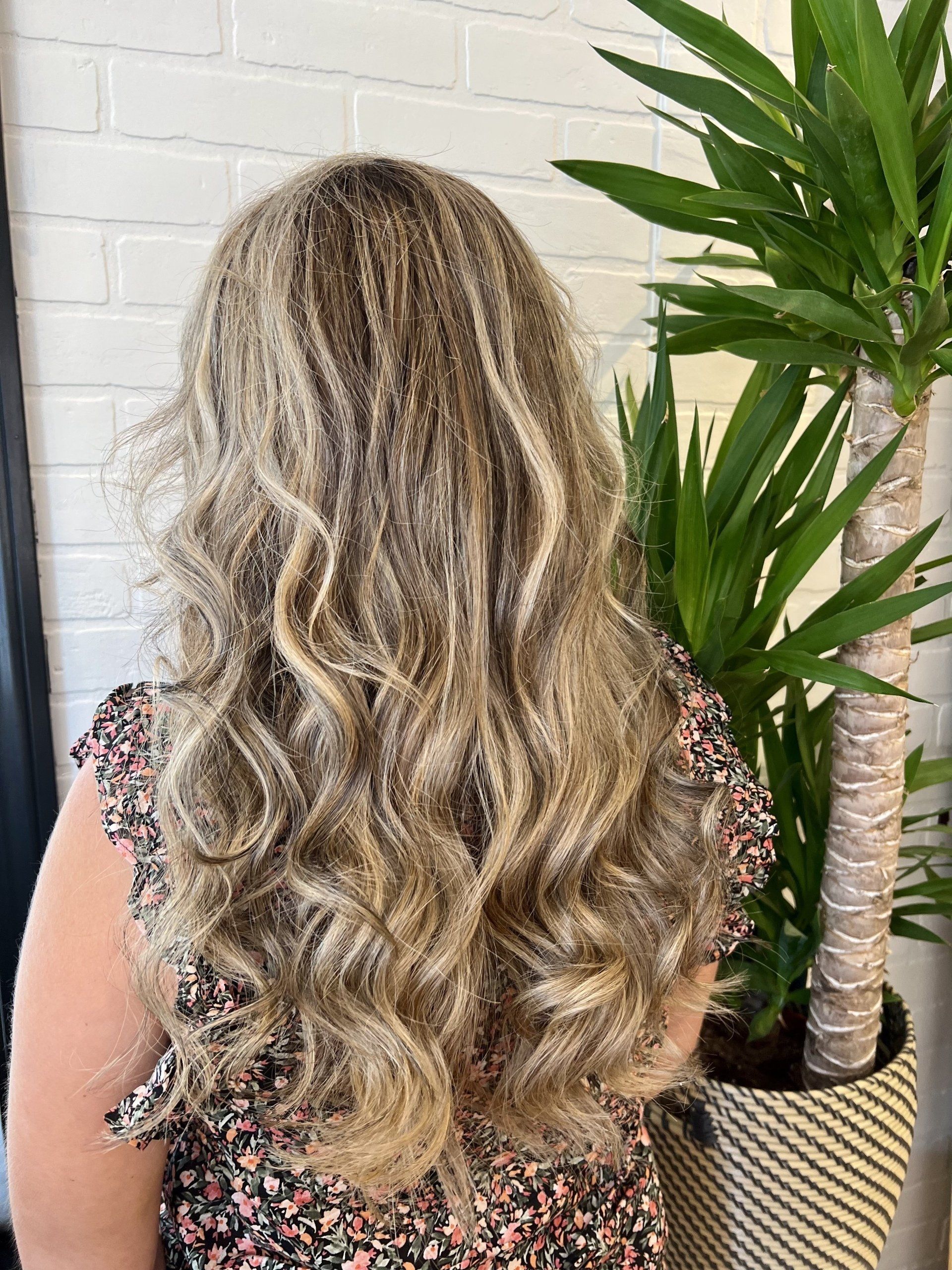 A woman with long blonde hair is standing in front of a potted plant.