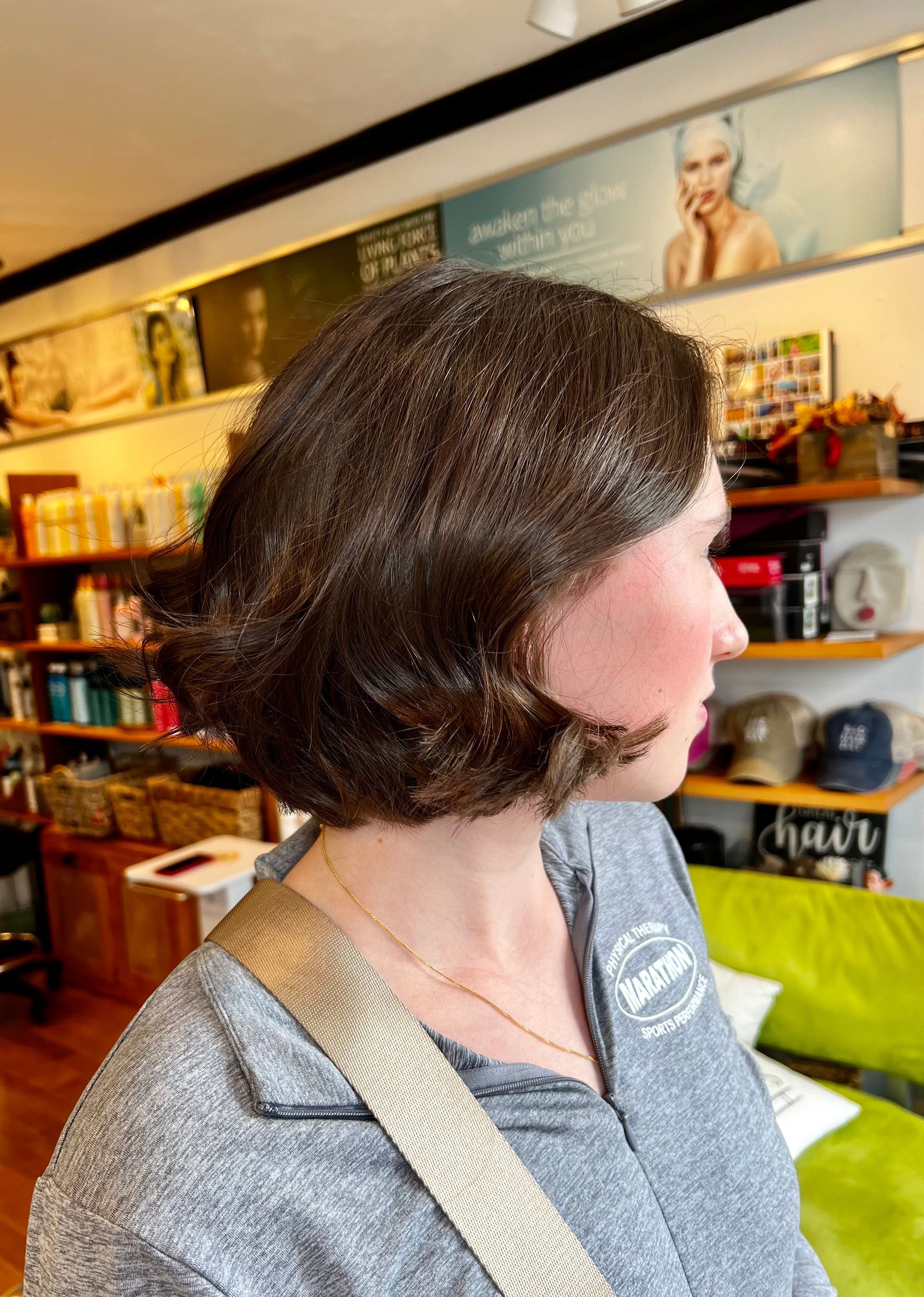 A woman with short brown hair is wearing a gray shirt and a gold strap.
