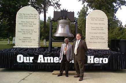 Two men are standing in front of a large bell and a sign that says our america hero