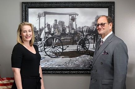 A man and a woman are standing in front of a framed picture of a horse drawn carriage.