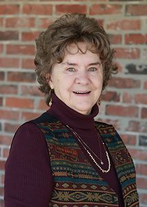 A woman wearing a turtleneck and a vest is smiling in front of a brick wall.