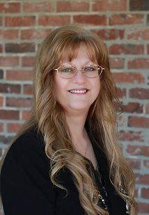 A woman wearing glasses and a black shirt is smiling in front of a brick wall.