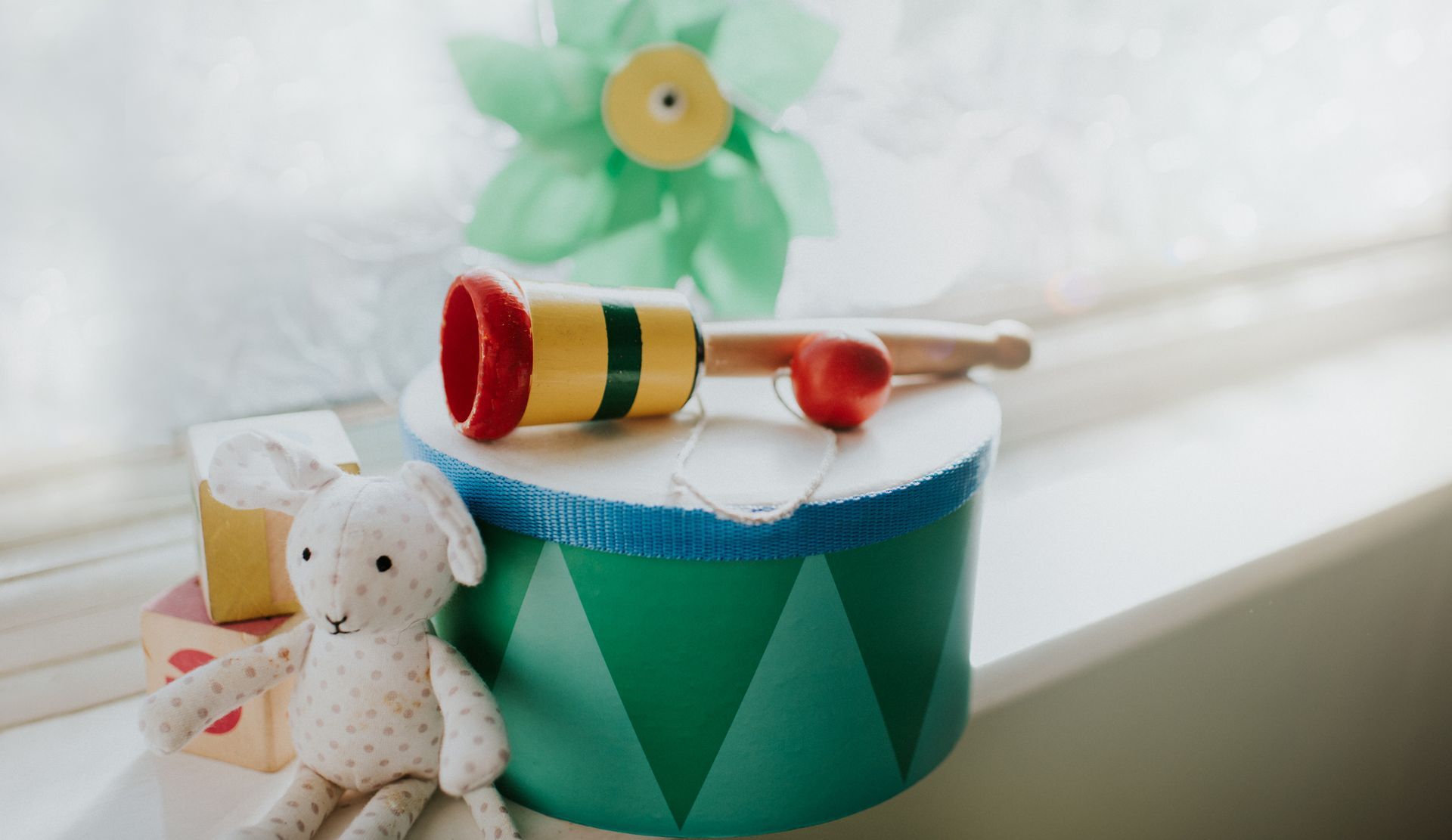 Tamburo giocattolo in legno con mazza, coniglietto e girandola sul davanzale di una finestra.