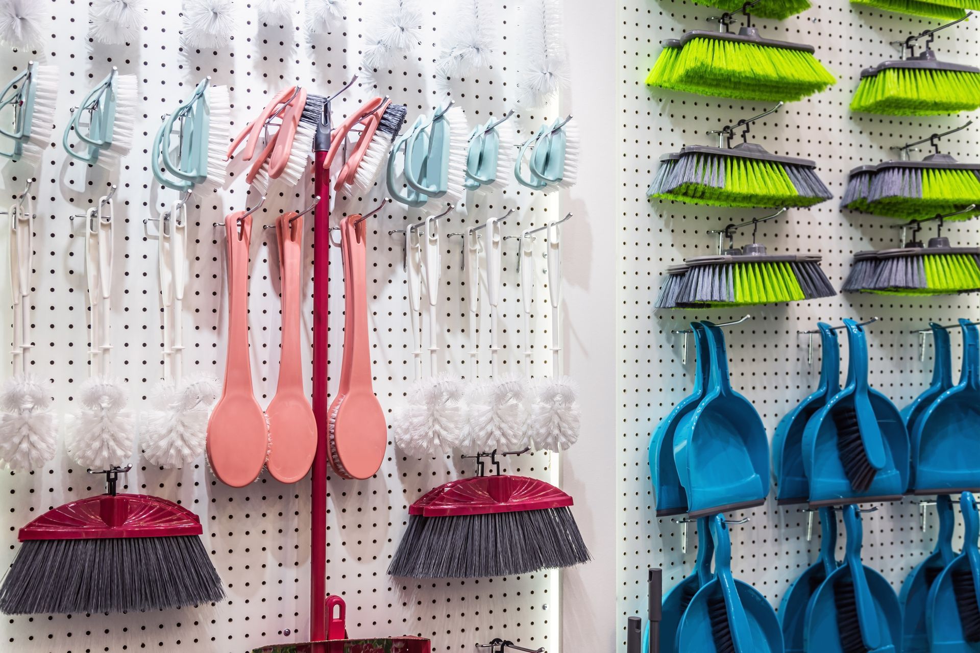Prodotti per la pulizia: spazzole, scope e palette appese a una parete forata in vari colori.