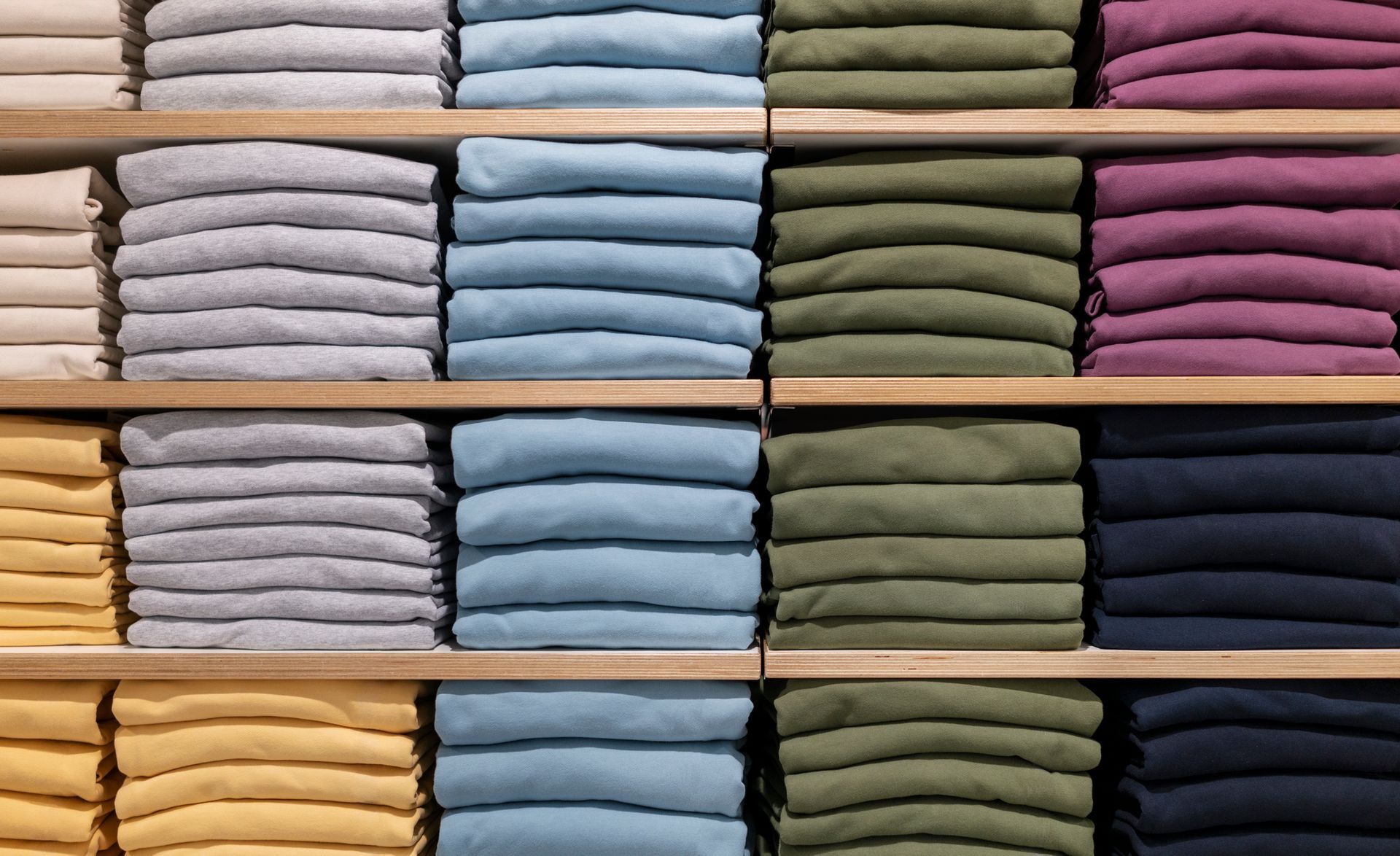 Scaffali pieni di vestiti colorati, ordinatamente piegati: crema, grigio, blu, verde, blu navy e viola.