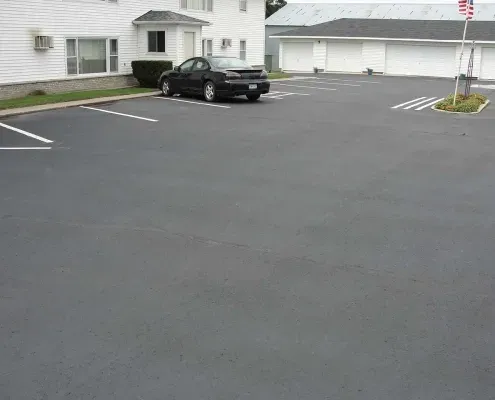 A black car is parked in a parking lot in front of a white house.