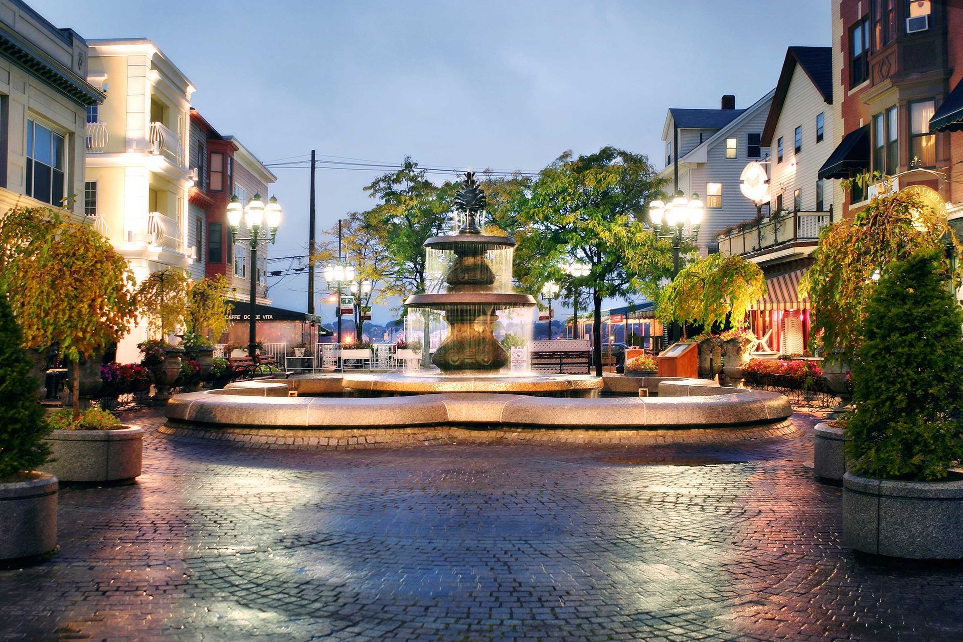 Evening view of a lit town square with a central fountain, wet cobblestones, and storefronts on both sides