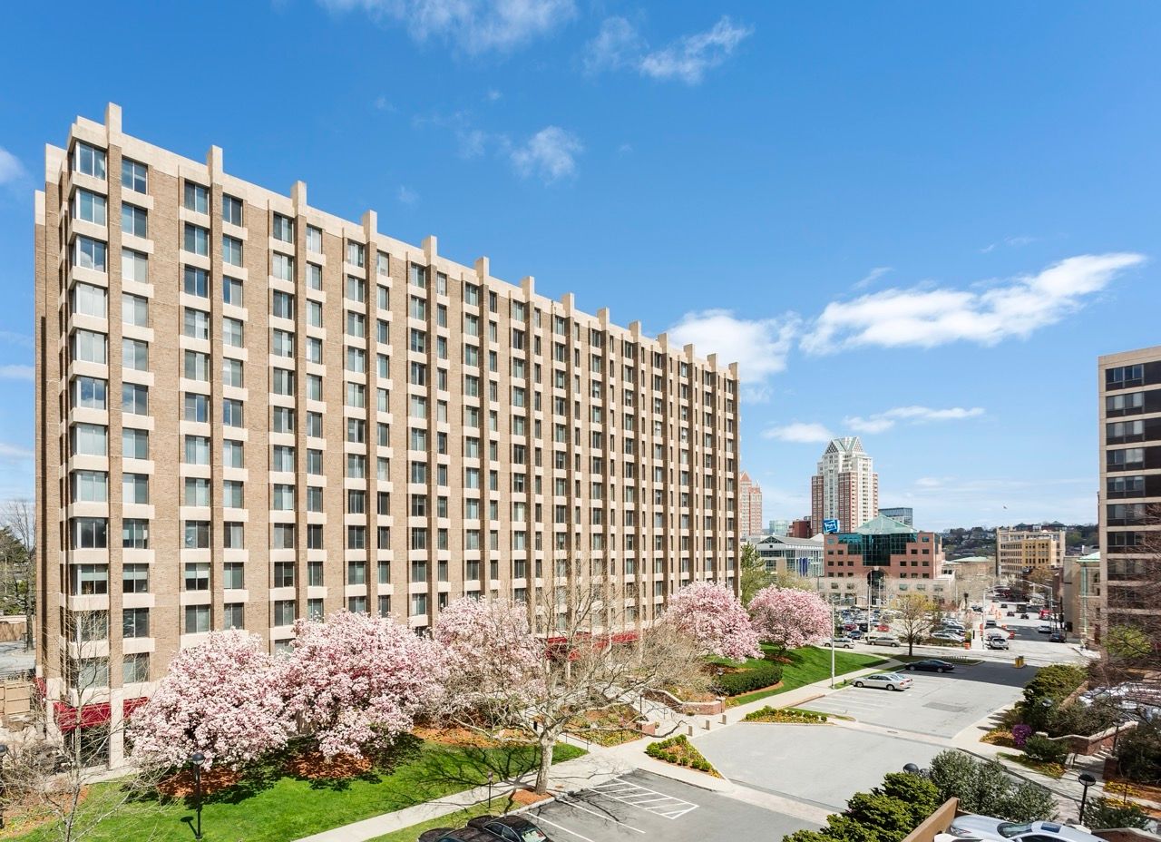 Exterior view of a multi-story brick apartment building with landscaped grounds and pink flowering trees.