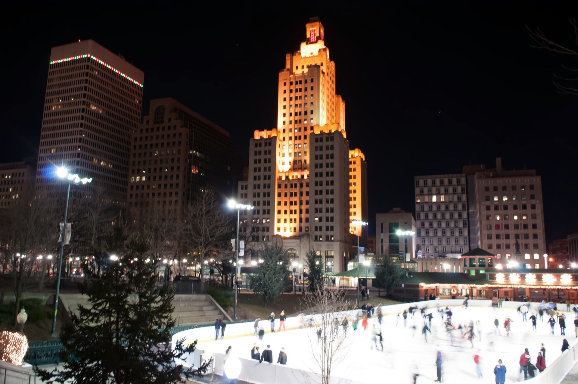 Nighttime city skyline with a brightly lit skating rink full of people