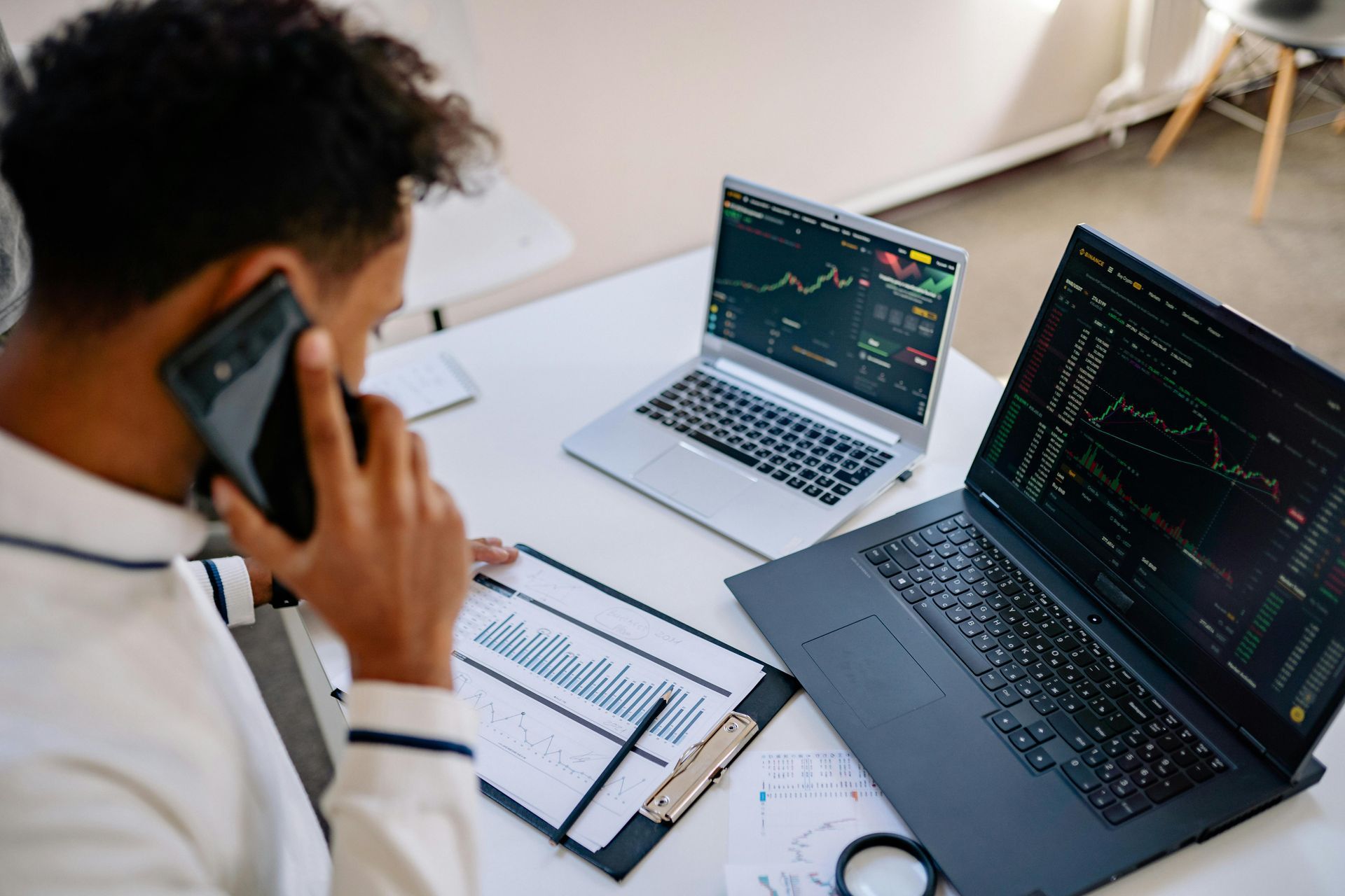Man on phone, analyzing stock charts on two laptops at a desk. Paper charts and pen are present.