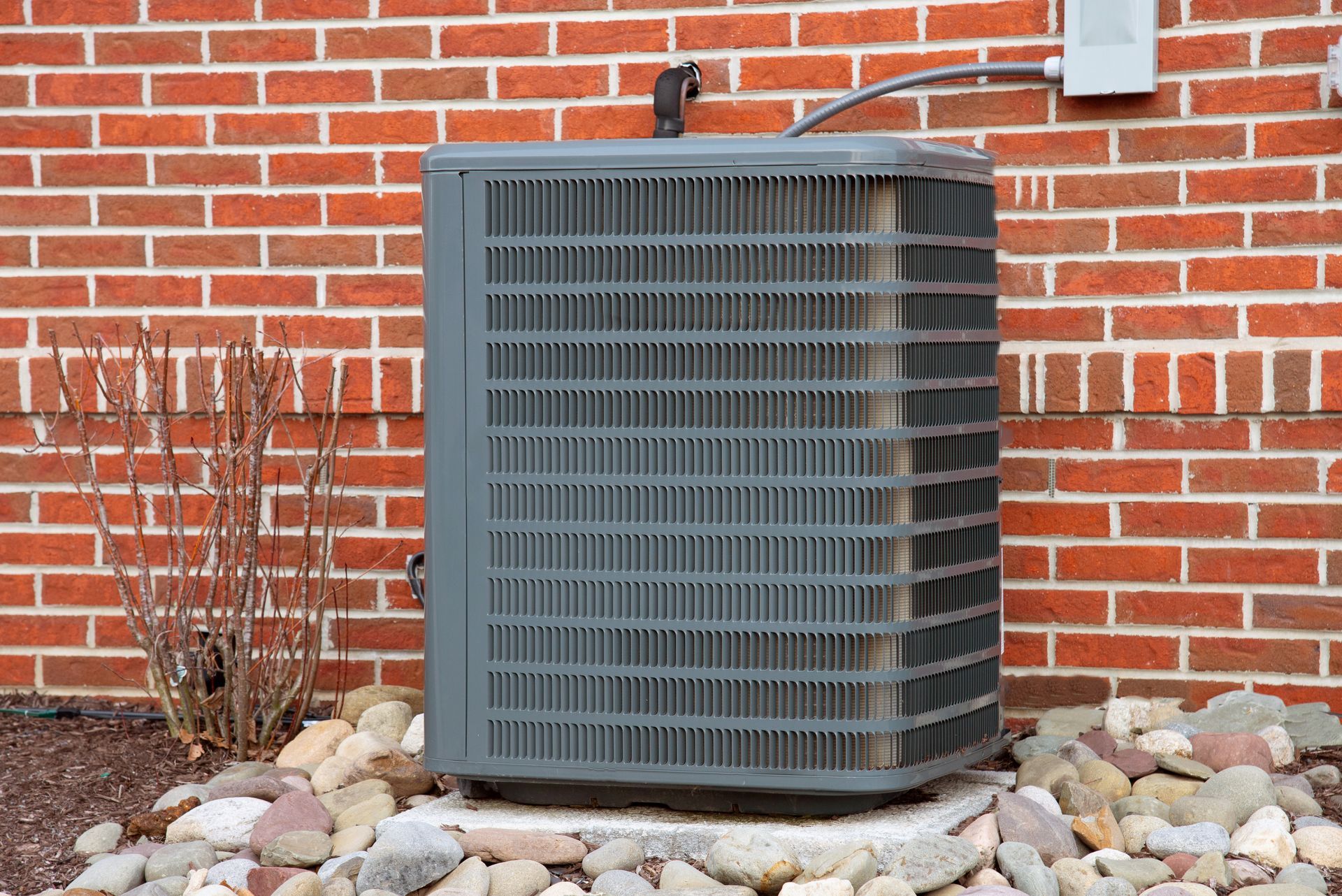 Air conditioning unit outside a brick building, sitting on a concrete pad with rocks.