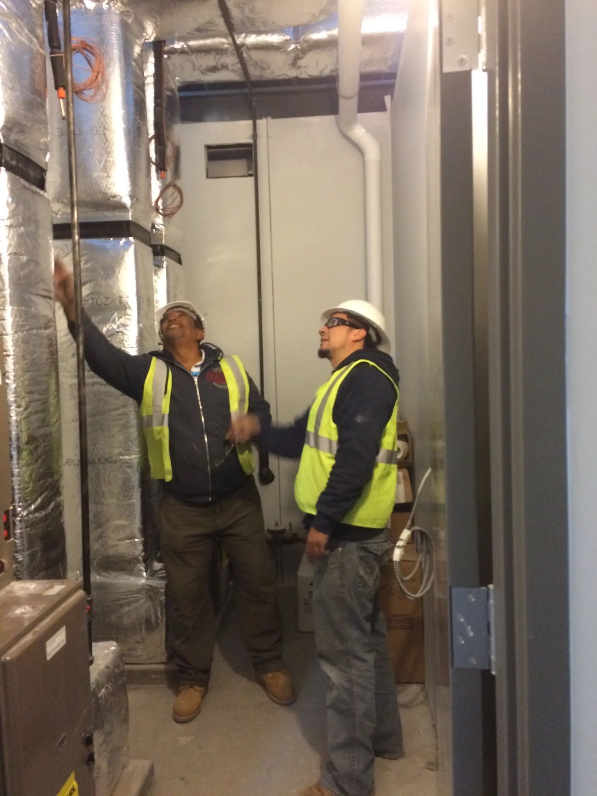 Two workers in safety vests examine pipes in an unfinished room.