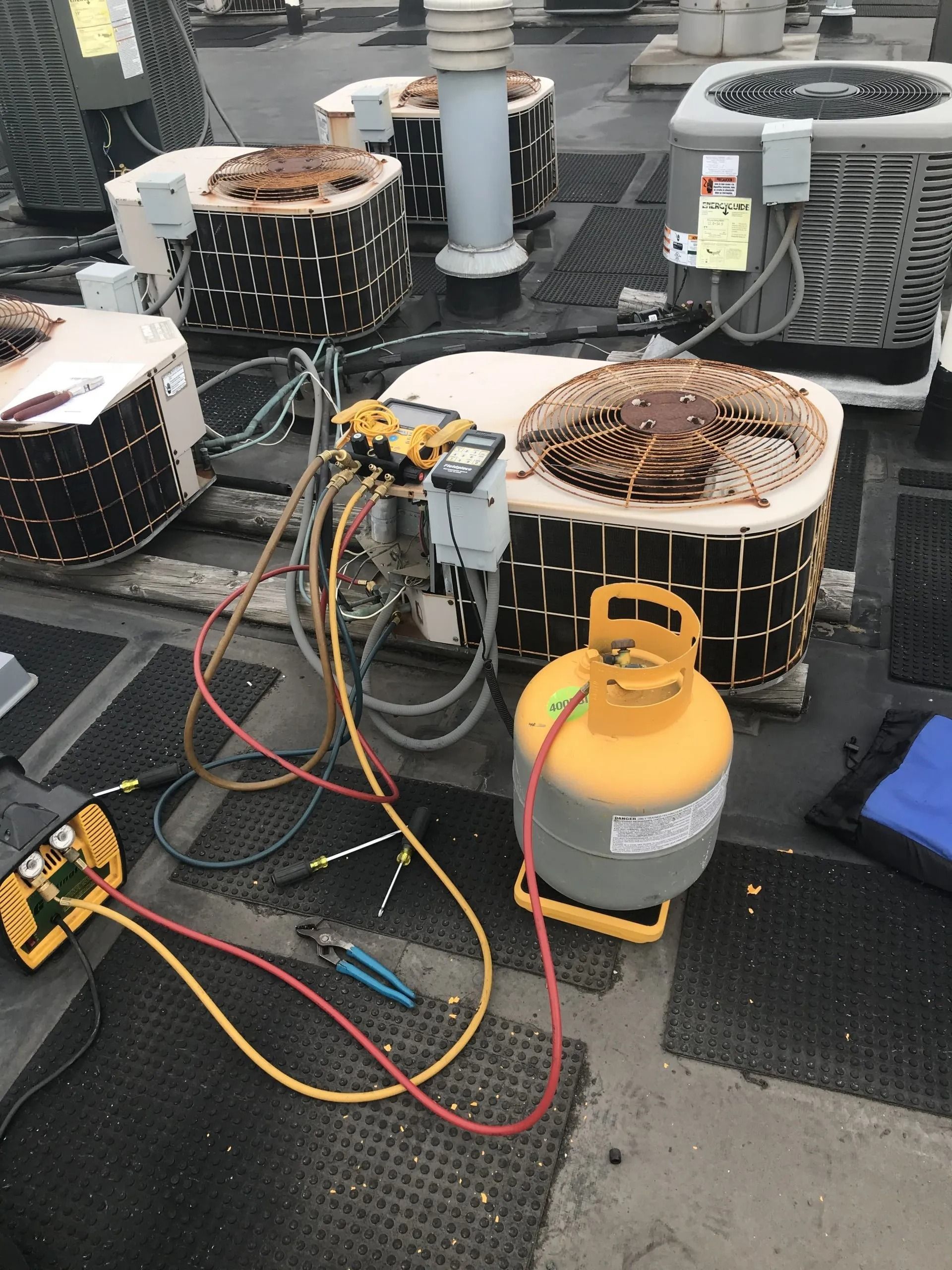 HVAC technician servicing rooftop air conditioning units. Yellow and red hoses connected to gauges and refrigerant tank.