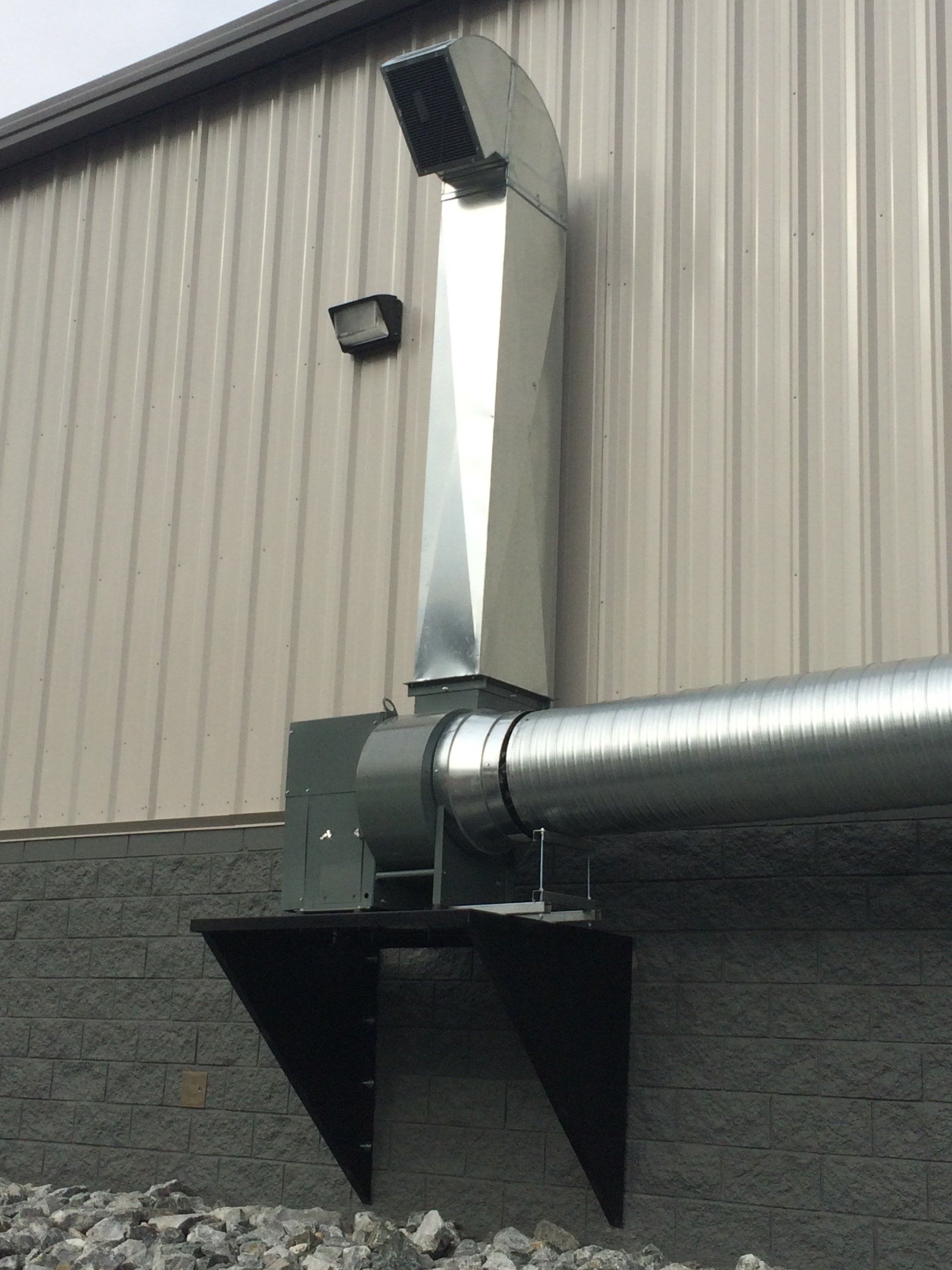 Silver exhaust fan mounted on a building's exterior wall, with a curved vent leading upwards.