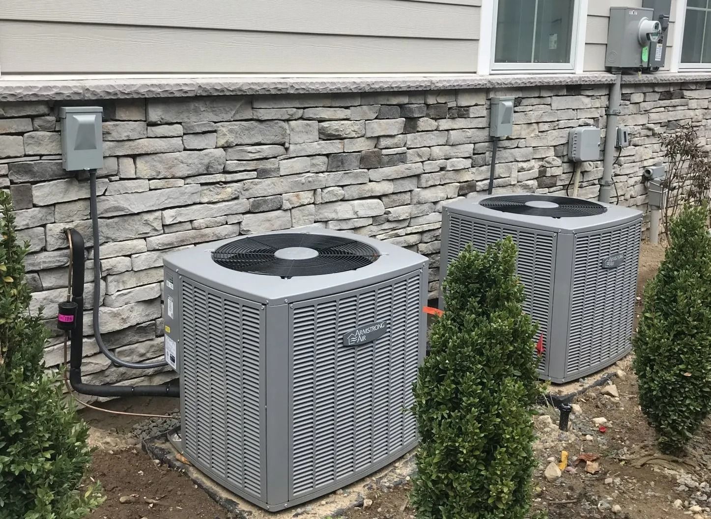 Two gray air conditioning units against a stone wall, near bushes and electrical boxes.
