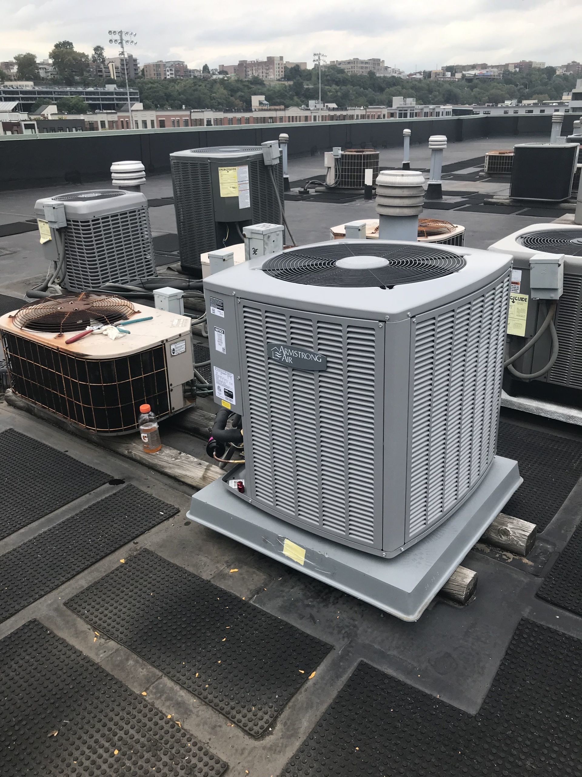 Several air conditioning units on a rooftop. Gray units, black roof with a city background.