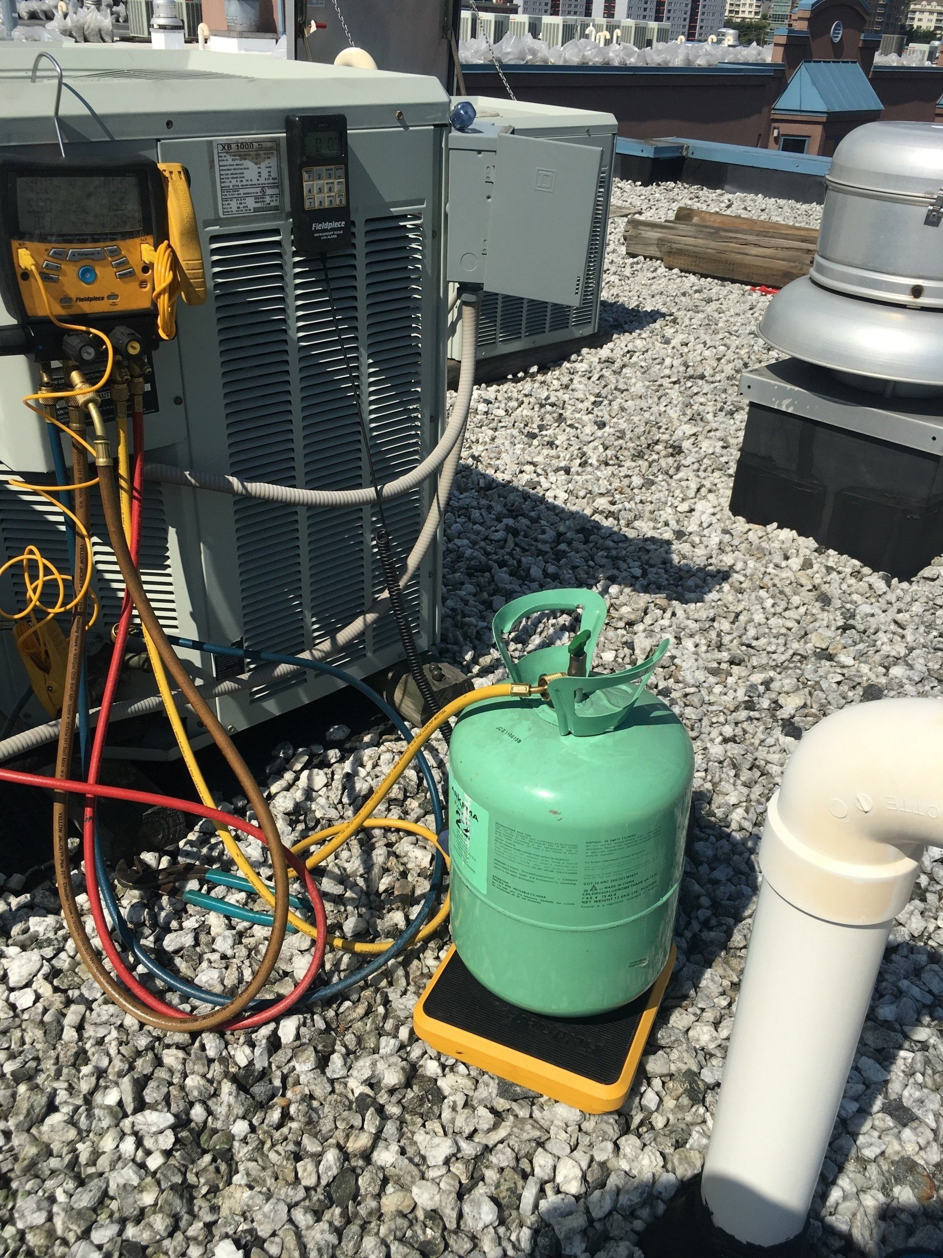 HVAC equipment on a rooftop, with gauges, hoses, and a refrigerant tank on a scale.