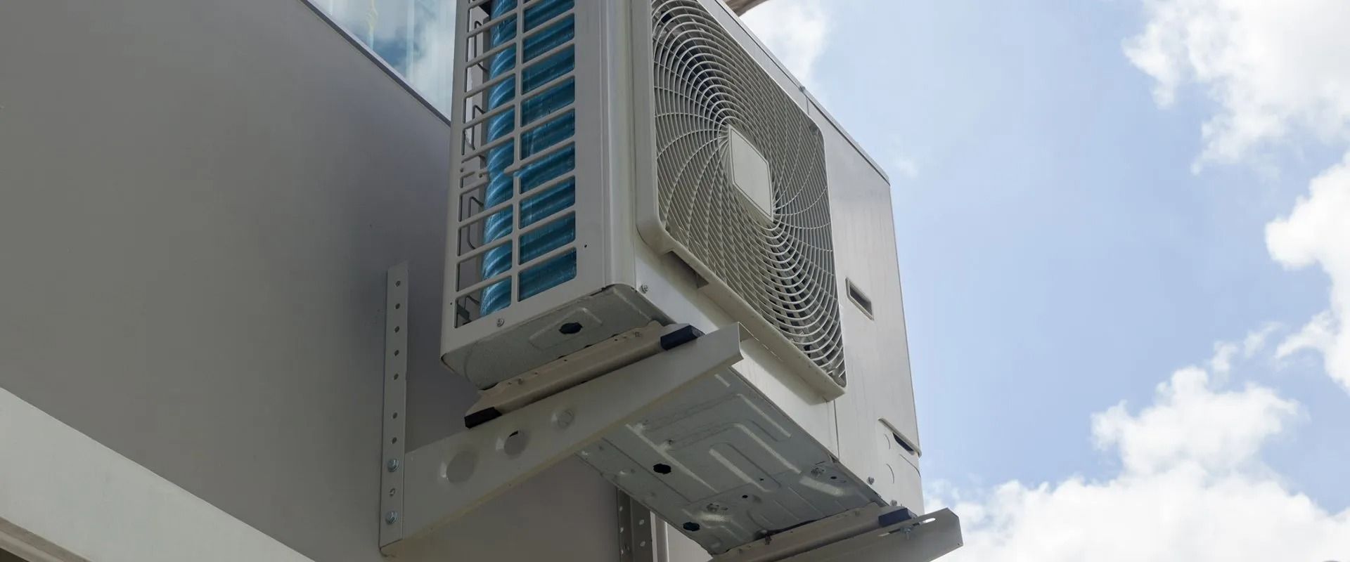 An air conditioning unit mounted on the side of a building, against a partly cloudy sky.