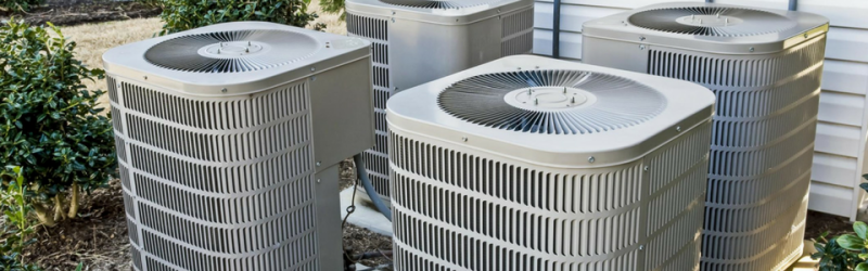 Four air conditioning units are in a yard, near greenery. Each has a fan on top.
