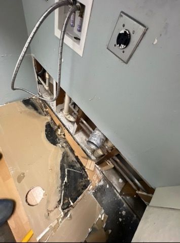 Damaged laundry room: ripped flooring, exposed pipes, electrical outlets, and water supply lines against a blue-gray wall.