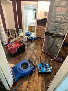 Flood damage in a room with drying equipment.  A washer is disassembled. Blue and red machinery are on the wet wooden floor.