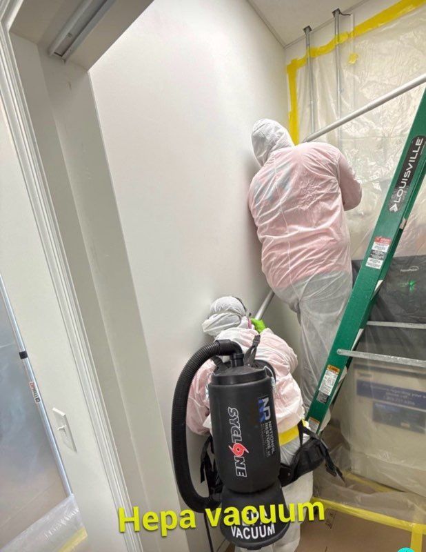Two workers in protective suits vacuuming a room. One on a ladder, using a Hepa vacuum.