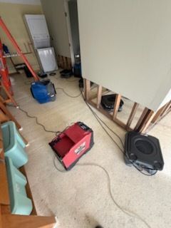 Dehumidifiers and fans drying a room; damaged wall exposed; orange ladder nearby.