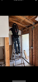 Person on a ladder, using a backpack vacuum in a room with exposed framing.