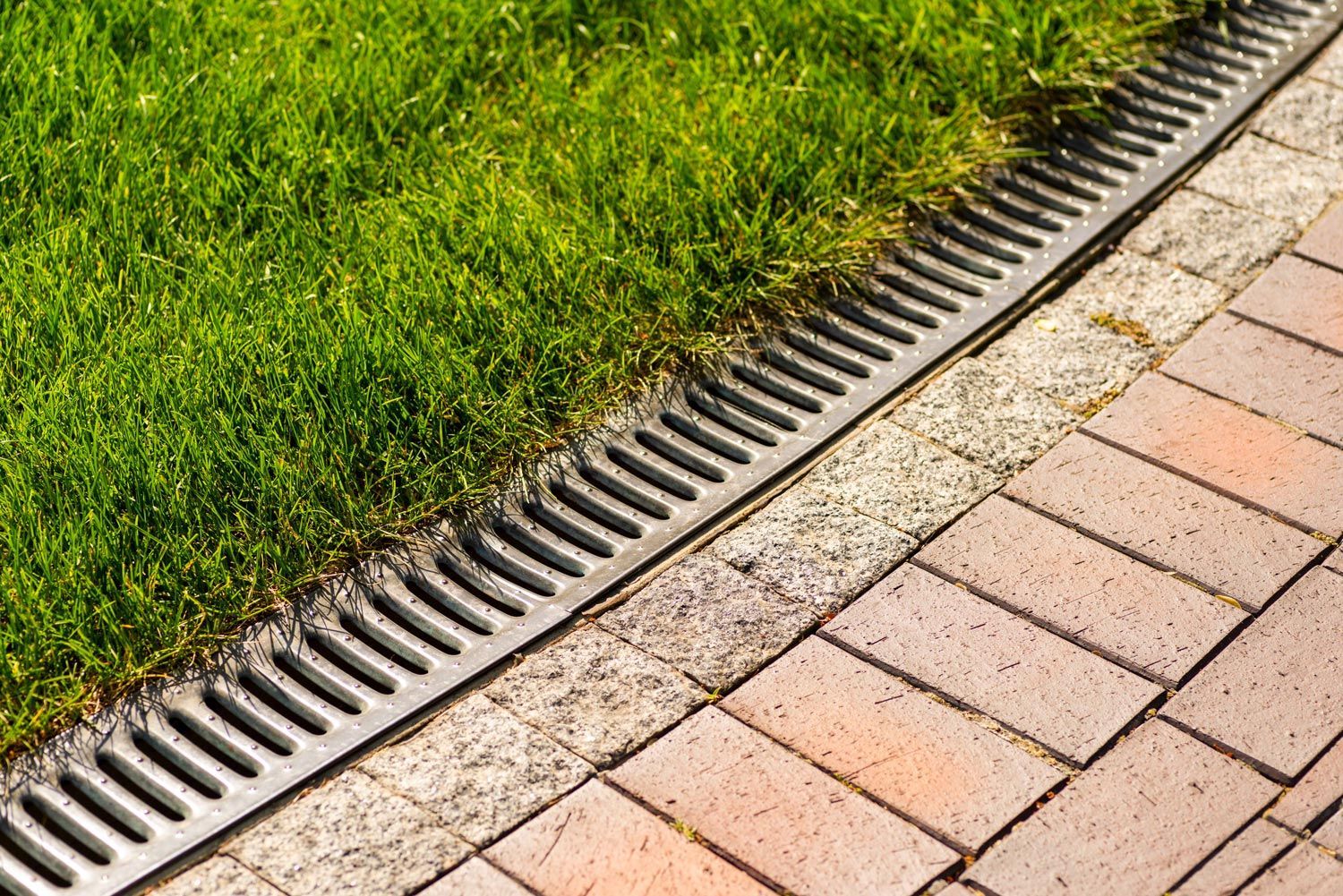 Grass beside a brick walkway with a black drainage grate along the edge
